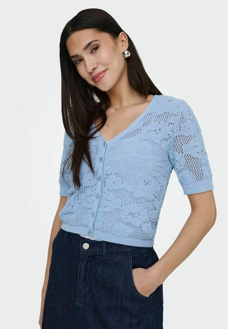 Femme aux longs cheveux noirs portant des boucles d'oreilles en argent et un cardigan en maille à manches courtes bleu clair avec motif et boutons, pantalon taille haute bleu foncé.