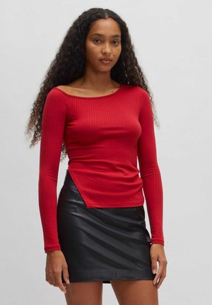 Rotes, geripptes Langarmoberteil mit einem schulterfreien Design und asymmetrischem Saum; kombiniert mit einem schwarzen Minirock aus Leder mit glatter Textur.