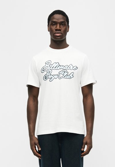 Άνδρας που φοράει λευκό μπλουζάκι με κοντά μανίκια και το κείμενο "Billionaire Boys Club", στέκεται μπροστά σε απλό ανοιχτό γκρι φόντο.