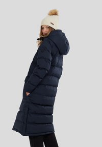 Dlouhá námořnická puffer bunda s kapucí, obsahující horizontální polstrování, postranní kapsy a zip. V kombinaci se smetanovou pletenou čepicí s pomponem.