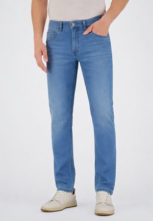 Uomo con jeans azzurri chiari e scarpe da ginnastica bianche, in piedi con una mano in tasca davanti a uno sfondo neutro.