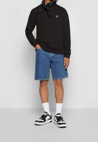 Felpa nera con logo CK, pantaloni shorts in denim e sneakers bianche e nere. L'outfit è completato da calzini bianchi e una sciarpa blu al collo.