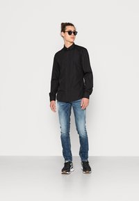Camicia nera a maniche lunghe, con bottoni, abbinata a jeans slim-fit blu chiaro. Sneakers nere con dettagli bianchi e color gomma. Accessorio: occhiali da sole neri.