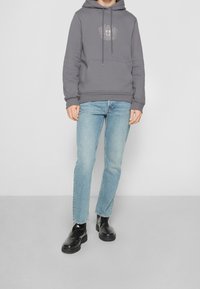 Felpa grigia con cappuccio, dotata di tasca frontale e logo, abbinata a jeans azzurri chiari e stivali neri alla caviglia con suola spessa.