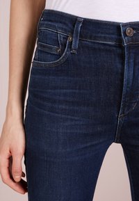 Tmavě modré džíny z denimového materiálu s přiléhavým střihem, klasickým pěti kapsovém designu, zapínáním na zip a kovovými detaily. Hladká textura.