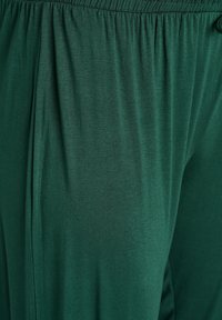 Zeleno, lahko tkanino z gladko teksturo, opremljeno z elastičnim pasom za udobje. Preprost dizajn, stranski naborki in brez vidnih okov.