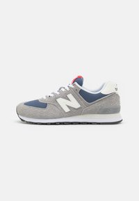 Zapatillas de ante gris y malla con acentos blancos, detalles azules y una etiqueta roja en la lengüeta. Cuenta con una suela de goma con diseño de dibujo.