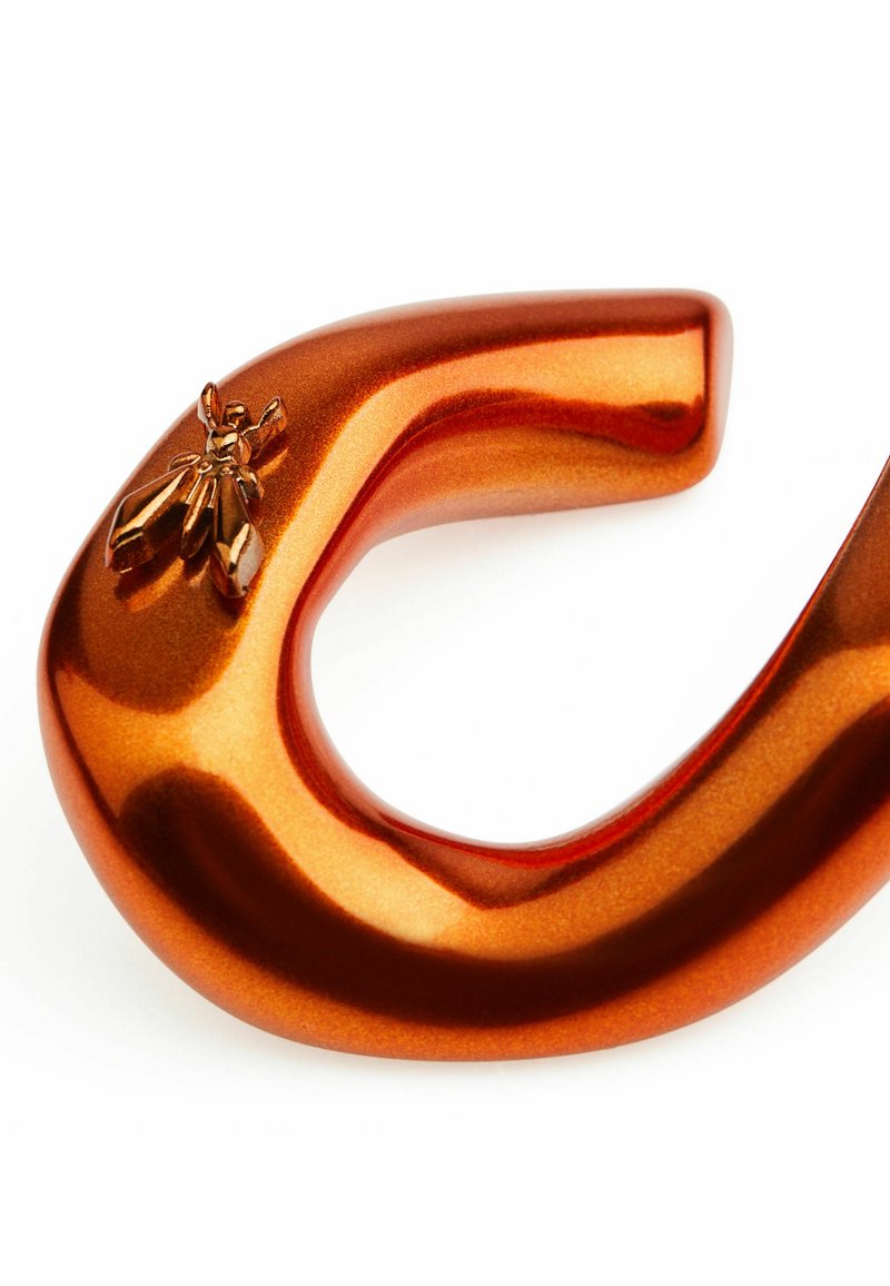 Oggetto curved di metallo arancione con una finitura lucida e un emblema di ape. Presenta una superficie liscia e una forma distintiva e fluida.