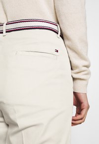 Pantaloni beige chiaro con cintura a strisce a contrasto nei colori navy, rosso e bianco. Il dettaglio della tasca posteriore include una piccola etichetta con il logo.