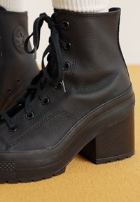 Black leather ankle boot with a high square heel, lace-up front, textured surface, and rubber sole. Features a circular logo on the side.