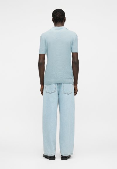 Άνδρας που στέκεται γυρισμένος με την πλάτη, φοράει ανοιχτό μπλε κοντομάνικο πόλο, φαρδιά τζιν παντελόνια και μαύρα παπούτσια πάνω σε λευκό φόντο.