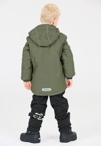 Giacca impermeabile verde con cappuccio e cuciture grigie, abbinata a pantaloni neri con dettagli del logo e robuste scarpe nere.