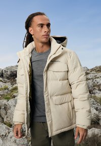 Man met dreadlocks in een beige Columbia pufferjack en een grijs shirt, staand op rotsachtig terrein onder een heldere blauwe lucht.