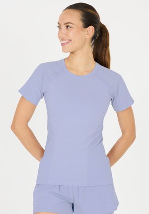 Frau mit Pferdeschwanz, die zur Seite lächelt, trägt ein eng anliegendes, kurzärmeliges, lavendelfarbenes Sportshirt und passende Shorts vor weißem Hintergrund.