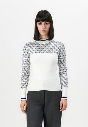 Donna con capelli neri lisci che indossa un maglione bianco aderente con maniche ornato e pantaloni grigio scuro, in piedi davanti a uno sfondo semplice.