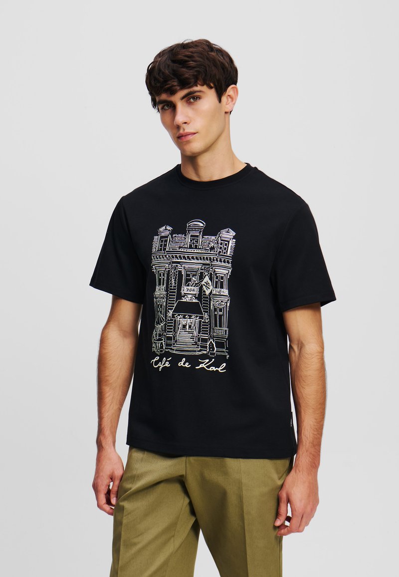 Juodas medvilninis marškinėliai su išsamia balta kontūro grafika, vaizduojančia kavinės pastatą, apačioje esantis tekstas "Café de Karl".