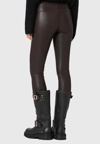 Brązowe skórzane legginsy w połączeniu z wysokimi czarnymi butami na obcasie z klamrami i teksturowanymi podeszwami, widok od tyłu na szarym tle.