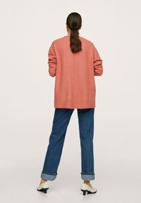 Różowy oversize'owy kardigan wykonany z miękkiej wełny, z prostym krojem, długimi rękawami i szwem na plecach, połączony z dżinsami w kolorze niebieskim z wywiniętymi nogawkami oraz białymi szpilkami.