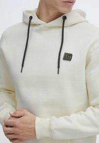 Felpa con cappuccio off-white con cordini neri e un piccolo logo patch verde e nero sul petto. Materiale morbido e polsini a costine.