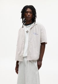 Camicia a maniche corte a righe beige e bianca, indossata sopra una maglietta bianca semplice, abbinata a pantaloni della tuta grigi chiari. Gioielli d'argento di rilievo.