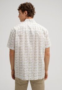 Kurzarmhemd aus leichtem Stoff mit weißem Hintergrund und beigem Blumenmuster; hat einen geraden Saum und einen Button-Down-Kragen.