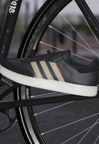 Czarny sneaker z beżowymi trzema paskami i czerwonym napisem "Velosamba". Posiada teksturowaną cholewkę i białą podeszwę, wyeksponowany na zębatce rowerowej.