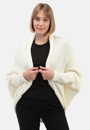 Pletený smetanový šál s ribovanou strukturou, otevřeným předním dílem a dlouhými rukávy, nošený přes černý top, s uvolněným, volným designem.