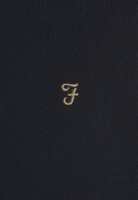 Zwarte stoffen shirt met een kleine gouden geborduurde letter "F" op de voorkant. Het materiaal lijkt glad met een fijne gebreide textuur.
