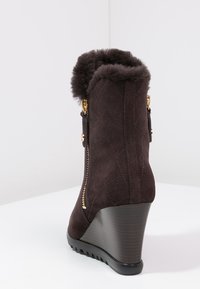 Brun kilklackad stövel i mocka med fuskpälskant, med guldfärgade dragkedjedetaljer och en strukturerad mörk träklack. Quiltad mönstring på skaftet.