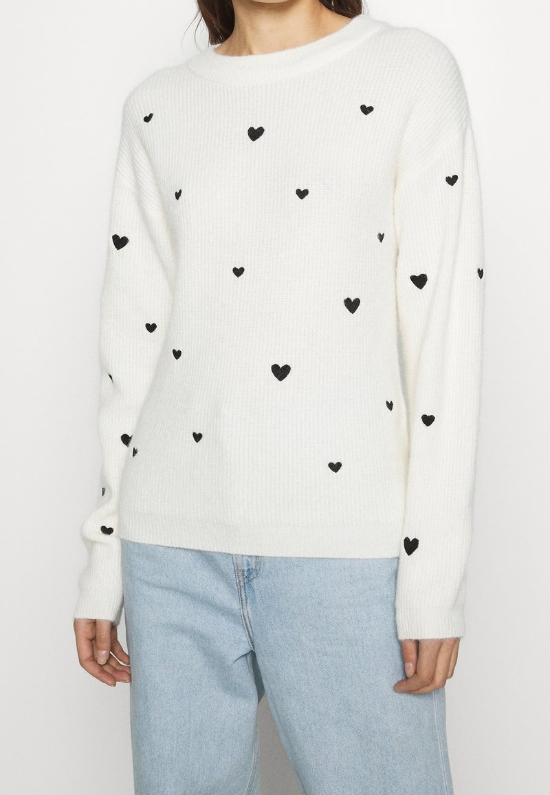 Femme portant un pull en maille blanc avec des motifs de cœurs noirs dispersés et un jean bleu clair, debout devant un fond uni.