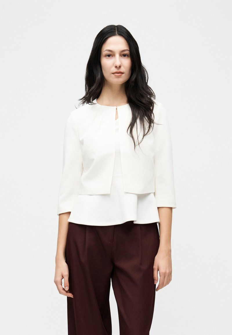 Femme aux longs cheveux bruns portant une veste courte blanche à manches trois-quarts sur un haut blanc, associée à un pantalon marron foncé taille haute.