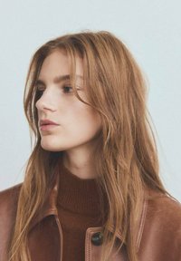 Veste en cuir marron à col montant, portée par-dessus un pull en maille marron. Cheveux longs et ondulés, sur un fond neutre.