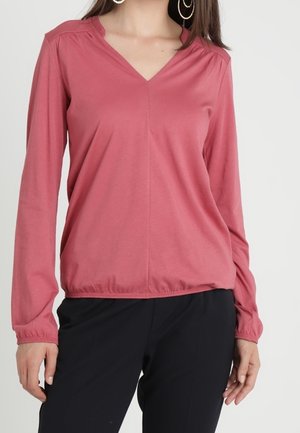 Kobieta ubrana w długim rękawem różową bluzkę z dekoltem w serek i elastycznym dołem oraz czarne spodnie, stojąca na jasnym, jednolitym tle.