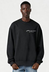 Jonge man die een zwart sweatshirt draagt met witte tekst "volg je hart" en een pijl, gecombineerd met lichtblauwe jeans, staand tegen een effen achtergrond.