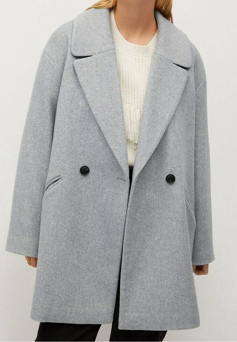 Kobieta w jasnoszarym, oversize’owym wełnianym płaszczu z szerokimi klapami, dwoma czarnymi guzikami i bocznymi kieszeniami, na kremowym swetrze z dzianiny.