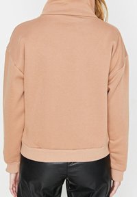 Persona con un suéter de cuello alto beige y pantalones de cuero negros, mostrada de espaldas contra un fondo liso.