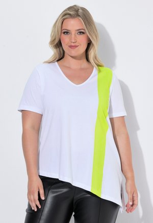 Vrouw met blond haar die een wit kortemouwen top draagt met een verticale gele streep en een zwarte leren broek, staand tegen een witte achtergrond.