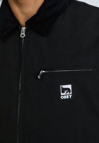 Casaco preto com colarinho de veludo cotelê, fecho frontal em zipper e um pequeno bolso no peito com zipper. Apresenta um patch com o logotipo "OBEY" no lado esquerdo.