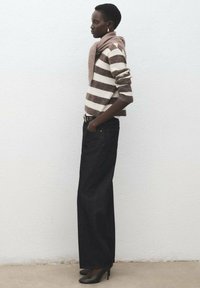 Suéter de lana a rayas en beige y marrón, combinado con jeans oscuros de tiro alto. La modelo lleva tacones negros elegantes y una bufanda colgando alrededor del cuello.