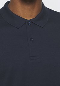 Mörkblå polo med ribbad krage, tvåknapplängd och en subtil söm under knapplängden. Lättviktig tyg.