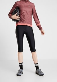 Personen har sorte cykelshorts, en blodrød jakke, sort og hvidt sokker, grå cykelsko og holder en sort hjelm under armen.