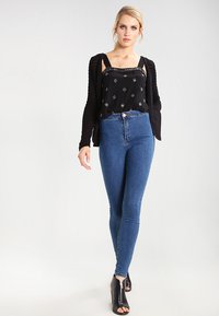 Svart ermeløs topp med hvite diamantmønstre, lagd under en strukturert svart cardigan, kombinert med høyhalsede blå jeans og svarte hæler.