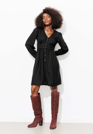 Robe noire à manches longues, col en V, fermeture à boutons, et taille froncée. Associée à des bottes bordeaux montantes et une texture lisse.