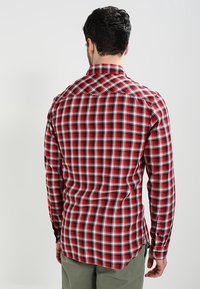 Mann in einem rot-weißen und schwarzen karierte Langarmhemd, von hinten gesehen, kombiniert mit olivgrünen Hosen vor einem schlichten Hintergrund.