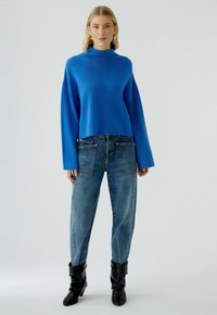 Blauer Langarmpullover mit hohem Kragen, kombiniert mit hellblauen Jeans mit aufgesetzten Taschen und schwarzen knielangen Stiefeln.