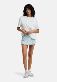Mujer con cabello rizado y oscuro, vestida con una camiseta blanca de gran tamaño, falda de mezclilla azul claro y zapatillas blancas de caña alta, de pie con las manos cerca de la cintura.
