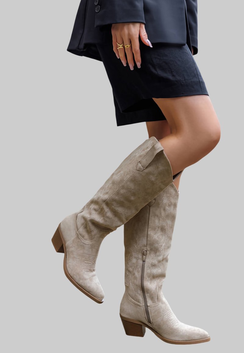 Bottes en daim beige montantes avec un bout pointu, un petit talon carré et une fermeture par zip latéral, présentant des détails de texture et de design subtils.