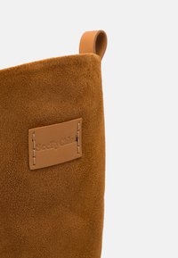 Sac cabas en daim marron avec une étiquette en cuir embossée du texte « SeeByChloé » et une bordure cousue. Poignée en cuir incurvée pour le transport.