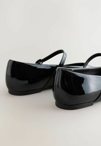 Bailarinas de charol negro con punta redonda, tacón bajo y correa ajustable. Superficie lisa y brillante con detalles de costura sutiles.