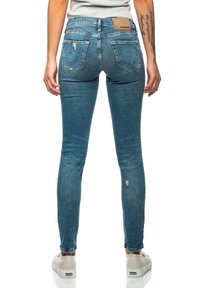 Smal blå denimjeans med en sliten yta, featuring ett beige logopatch på midjan och två bakfickor. Bärs med ljusa skor.